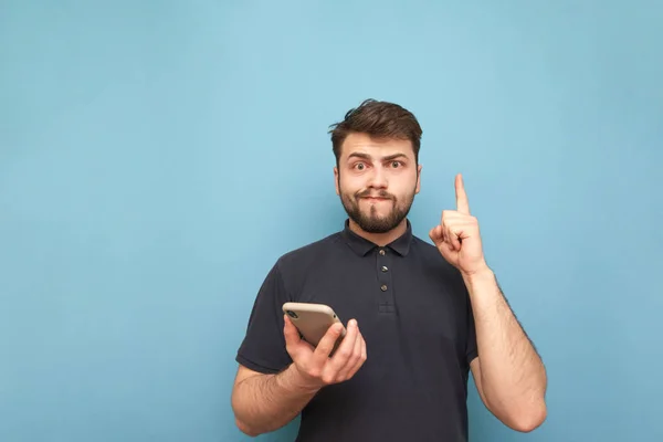 Portrait of a funny man with a beard and a phone in his hand on a blue background, looking into the camera and making a funny face. Confused man with a smartphone shows an empty space. Isolated.