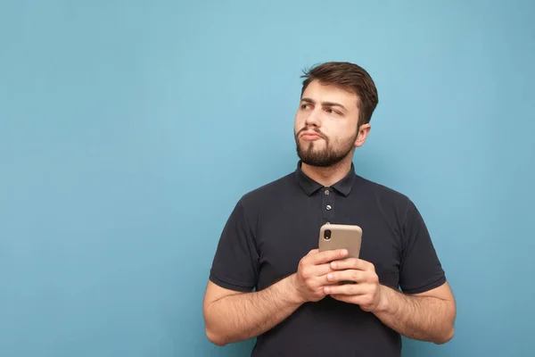 Retrato de un hombre desconcertado en la ropa casual se encuentra con un teléfono inteligente en sus manos, mirando pensativamente de lado sobre un fondo azul. Espacio de copia — Foto de Stock