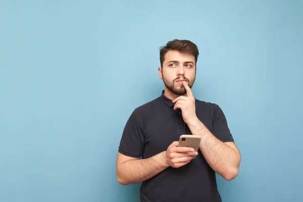 Thoughtful man with a beard standing on a blue background with a smartphone in his hand, looking sideways and thinking wearing a dark t-shirt. Isolated. Copy space