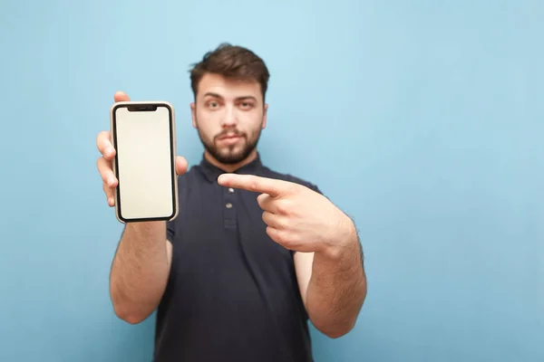 Smartphone με κενή λευκή οθόνη στα χέρια ενός ενήλικα άντρα με μούσι, φορώντας ένα σκούρο πουκάμισο και στέκεται σε μπλε φόντο. Ο άνθρωπος κρατά ένα μοντέρνο τηλέφωνο στα χέρια του, εστίαση smartphone. Απομονωμένες — Φωτογραφία Αρχείου
