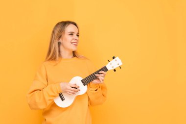 Elinde ukulele ile Mutlu kız turuncu bir arka plan üzerinde duruyor, yana bakar ve gülümsüyor, sarı rahat giysiler giyer. Gülümseyen kız müzisyen ukulele çalar. Izole.