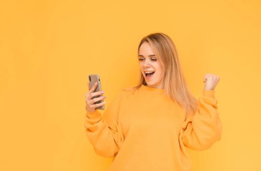 Gülümseyen sarışın kız parlak giysiler içinde bir akıllı telefon kullanır ve sarı bir arka plan üzerinde sevinir. Mutlu bir kız portresi bir akıllı telefon ekranına bakarak ve zafer sevinir, izole.