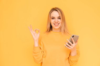 Sarı bir arka plan üzerinde elinde bir akıllı telefon ile parlak giysiler içinde Mutlu kız, parmaklarıyla bir Ok jest gösterir ve kameraya bakar. Gülümseyen sarışın akıllı telefonda internet kullanıyor