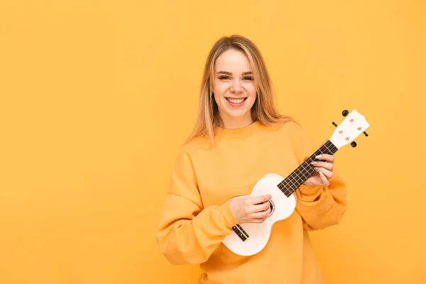 Ritratto di una ragazza sorridente in abiti vivaci, in piedi con una chitarra hawaiana tra le mani su uno sfondo giallo, guardando con fotocamera e gioendo. Musicista bionda suona ukulele e ride, isolato . — Foto Stock