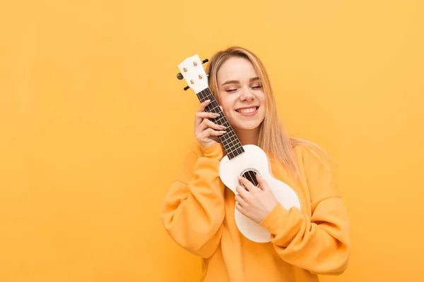 Menina bonito abraça ukulele com os olhos fechados e sorri, isolado em um fundo amarelo, vestindo roupas laranja. Retrato de uma menina feliz com guitarra havaiana nas mãos. Espaço de cópia — Fotografia de Stock