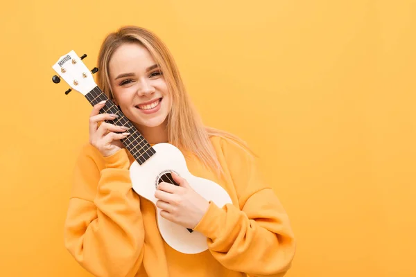 Glimlachend blondine in gele kleren met ukulele in de hand wordt geïsoleerd op een oranje achtergrond, kijkend in de camera en blijdschap. Portret van een positief meisje met Hawaiiaanse gitaar in de hand. Ruimte kopiëren — Stockfoto