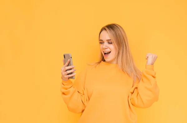 Uma menina loira sorridente usa um smartphone em roupas brilhantes e se alegra em um fundo amarelo. Retrato de uma menina feliz olhando para a tela de um smartphone e se alegra na vitória, isolado . — Fotografia de Stock