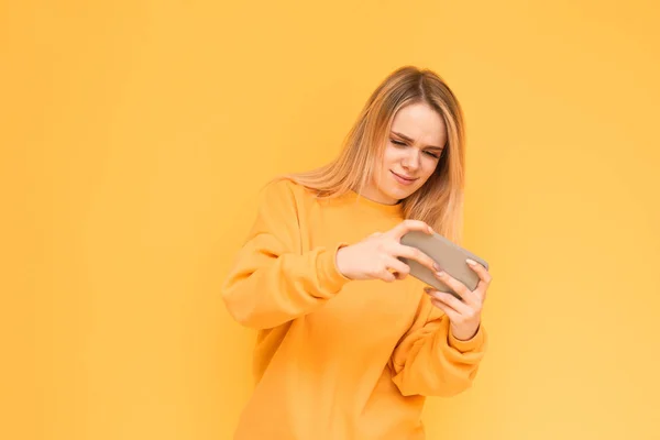 Menina concentrada em roupas amarelas jogando videogames em um smartphone, focada em olhar para a tela. Jogador loiro joga jogos móveis em um fundo amarelo. Espaço de cópia — Fotografia de Stock