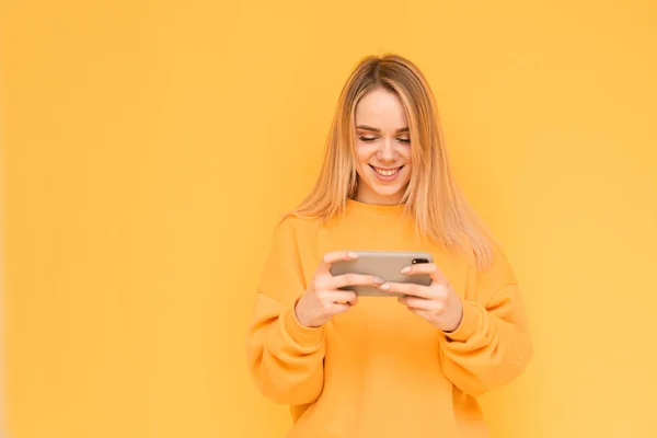 Портрет усміхненої дівчини в яскраво-помаранчевому одязі з смартфоном в руках на жовтому тлі, дивлячись на екран і посміхаючись. Щаслива дівчина грає в мобільні ігри на смартфоні . — стокове фото