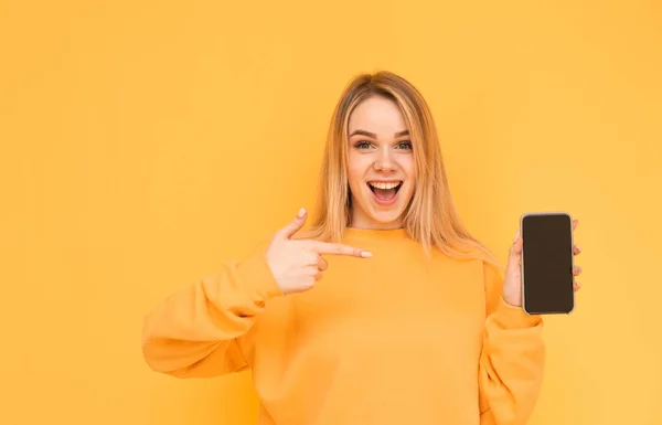 Menina surpresa em roupas laranja está em um fundo amarelo com um smartphone em sua mão, mostra um dedo em uma tela em branco preto, olha para a câmera com um rosto chocante. Isolados . — Fotografia de Stock