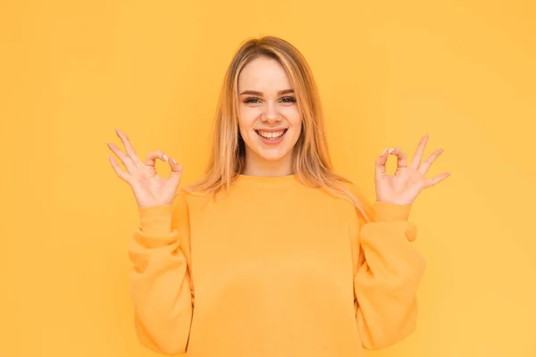 Retrato de uma menina positiva em roupas laranja em pé sobre um fundo amarelo, olhando para a câmera, sorrindo e mostrando o sinal OK com as varas.A menina está bem, ela está satisfeita. Isolados — Fotografia de Stock