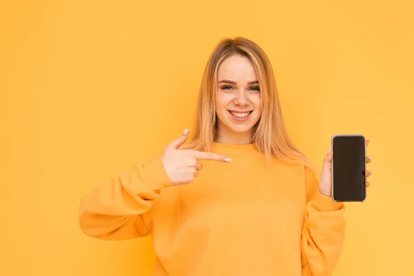 Happy girl in bright clothes stands on a yellow background with a smartphone in her hand, shows her finger on the screen, looks into the camera and smiles. Isolated. — Stock Photo, Image