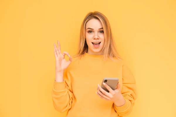 Menina loira doce em roupas laranja fica com um smartphone na mão em um fundo amarelo, mostra aos dedos um sinal ok e olha para a câmera com um rosto surpreso. Isolados . — Fotografia de Stock