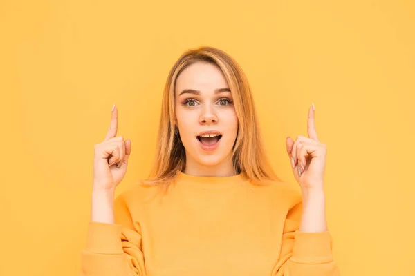 Attractive blonde with a shocking face is isolated on a yellow background, looks at the camera with astonishment and shows fingers up. Surprised girl shows her fingers in an empty place. Copy space
