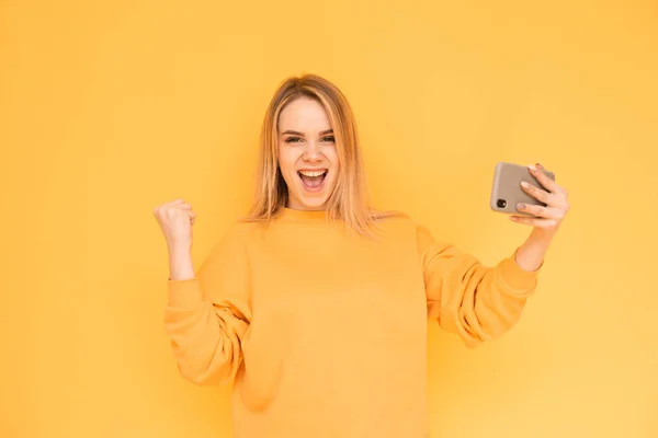 Menina loira alegre em uma roupa casual brilhante segura um smartphone em suas mãos, olha para a câmera e grita. Menina adolescente sorridente com um telefone em seus braços se alegra em um fundo amarelo. A menina — Fotografia de Stock