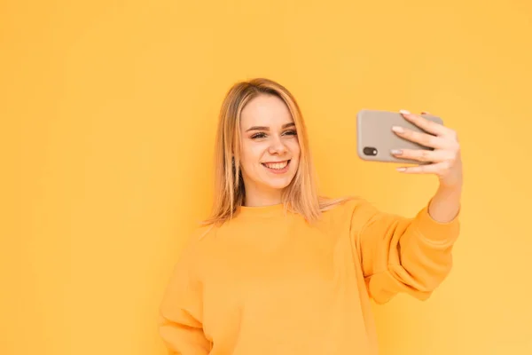 Привлекательная юная леди в яркой одежде делает селфи на желтом фоне, позируя для камеры смартфона, улыбаясь и глядя в телефон. Изолирована. Копирование пространства — стоковое фото