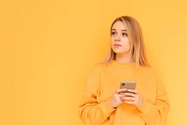 Menina atraente em roupas brilhantes está de pé sobre um fundo amarelo com um smartphone na mão, olhando para o lado com um rosto sério. Retrato de uma loira pensativa olha para o lado e pensa . — Fotografia de Stock