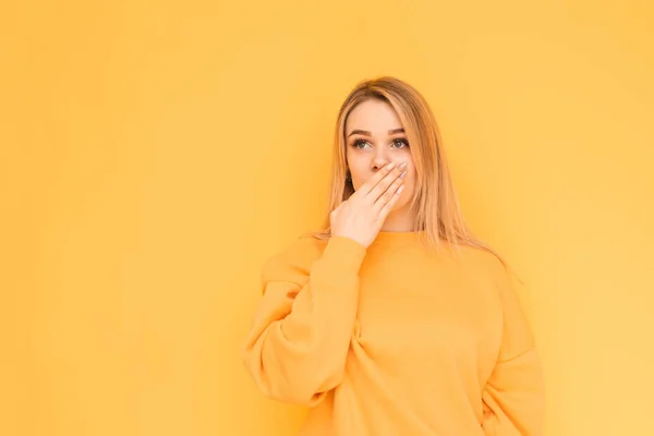 Surprised girl in orange clothes stands on a yellow background, looking sideways, shook hands with her face. Isolated Copy space