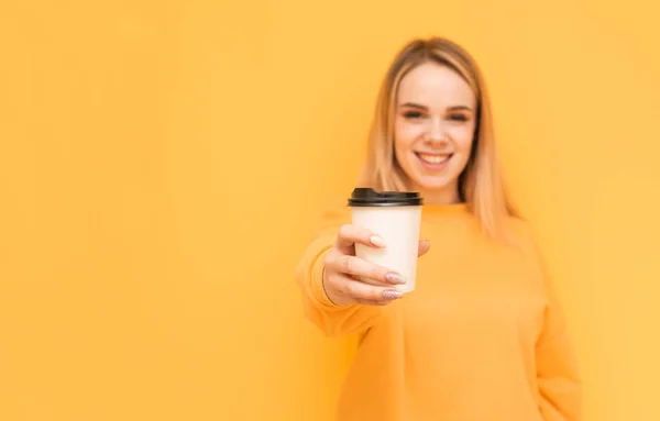 Улыбающаяся блондинка в оранжевом свитере показывает чашку кофе в камере на желтом фоне, глядя в камеру. Сосредоточьтесь на чашке кофе — стоковое фото
