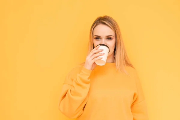 Jolie fille dans un pull orange se tient sur le fond d'un mur jaune, boit du café dans une tasse en papier, sourit et regarde la caméra. Isolé . — Photo