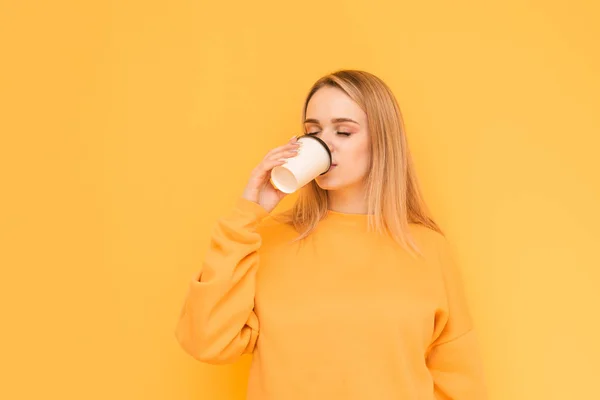 Douce dame dans un pull orange boit du café dans une tasse en papier avec les yeux fermés sur un fond jaune. Jeune amateur de café boit du café, obtient plaisir. Isolé . — Photo