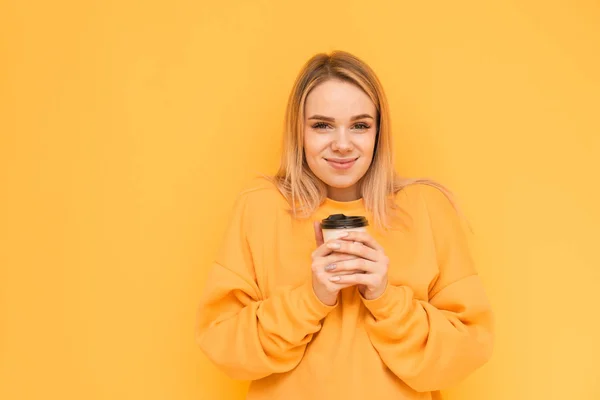 Девушка в оранжевом свитере стоит на желтом фоне с чашкой кофе в руке, смотрит в камеру и мило улыбается. Изолированная девочка любит кофе . — стоковое фото