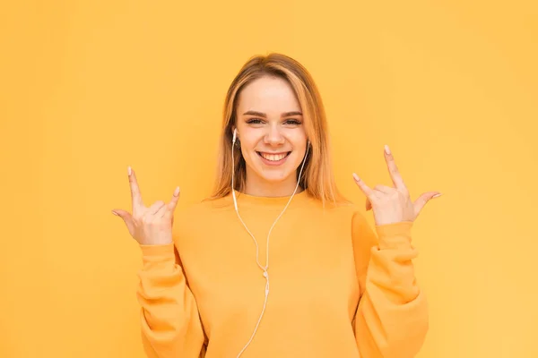 Ritratto di una ragazza sorridente con le cuffie su sfondo giallo che indossa un maglione arancione, guarda nella macchina fotografica e sorridente.Ragazza in cuffia ascolta la sua musica preferita e gioisce . — Foto Stock