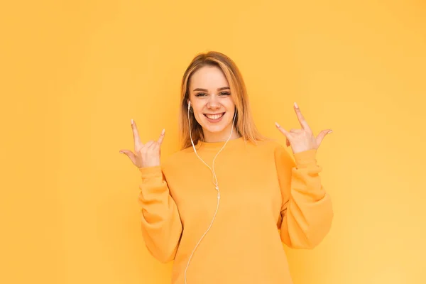 Happy girl listens to music in the headphones on a yellow background, shows a heavy metal tin, looks into the camera and smiles. Lady listens to her favorite music in her headphones, happy. Isolated