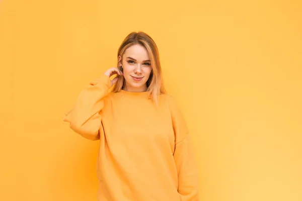 Modelo atraente em uma camisola laranja posa, sorri e olha para a câmera, isolada em um fundo amarelo. Senhora positiva fica no fundo de uma parede amarela, sorrisos bonitos . — Fotografia de Stock