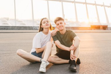 Güzel bir gün batımı arka planda oturan şık bir Casual giyim Charming genç çift, kamera üzerinde poz ve gülümseyen. Güzel bir arka plan ile akşam rahatlatıcı romantik mutlu çift