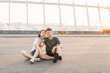 Gün batımı ve bir akıllı telefonda fotoğraf yapma bir şehir manzara arka planda yerde oturan mutlu çift. Şık bir Casual giyim gülümseyen çift selfie yapar. Tatlı çift rahatlatıcı