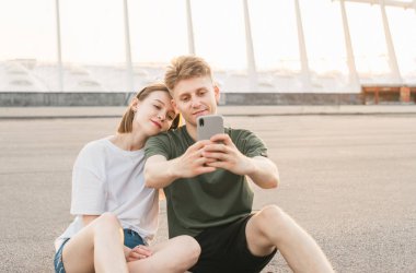 Güzel genç çift kare üzerinde oturan kentsel arka plan üzerinde selfie almak, kamera bakarak ve akıllı telefon gülümseyen. Şık genç adam ve kız Smartphone bir fotoğraf çekmek. Yaşam tarzı.