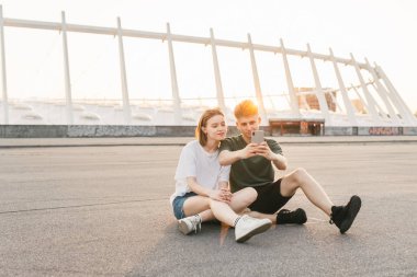 Şık kıyafetler kaldırım üzerinde oturan sevimli çift, günbatımı güzel kentsel manzara ve bir akıllı telefon kullanarak. Sokakta genç adam ve kız dinlenme, yere oturmak ve fotoğraf yapmak.