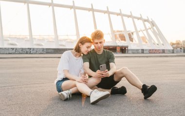 Güzel çift gün batımı ve kullanım Smartphone güzel bir şehir manzara arka planda rahatlatıcı, kaldırım üzerinde oturan, ekranda bakıyor. Elle telefon ile genç çift, kentsel arka plan