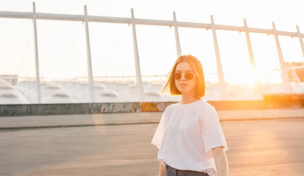 Retrato de menina atraente em camiseta branca e óculos de sol no fundo da paisagem da cidade no pôr do sol. Mulher bonita elegante no fundo de um belo pôr do sol . — Fotografia de Stock