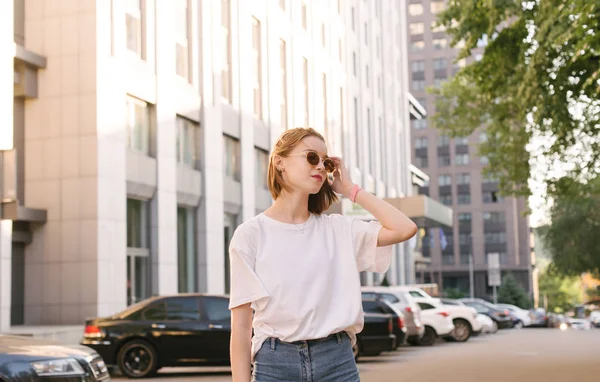 Ritratto di strada di una ragazza elegante in abiti casual per una passeggiata in una calda giornata estiva. Attraente ragazza in occhiali da sole posa sulla strada sullo sfondo del paesaggio urbano . — Foto Stock