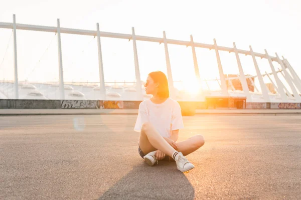 Stylisches Mädchen sitzt auf der Straße vor dem Hintergrund eines schönen Sonnenuntergangs und moderner Architektur an einem warmen Sommerabend und blickt aus dem Weg. Sommerkonzept. — Stockfoto