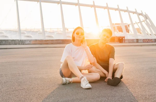 Beautiful young couple sitting on the background of an incredible sunset, looking into the camera and smiling. Wear a stylish casual clothing. Couple posing on the background of the sun at sunset. — Stock Photo, Image