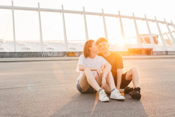 Estilo de vida foto de jovem casal sentado ao pôr do sol e abraço, menina beija menino, jovem olhando para a câmera. Retrato urbano de um casal amoroso ao pôr do sol . — Fotografia de Stock