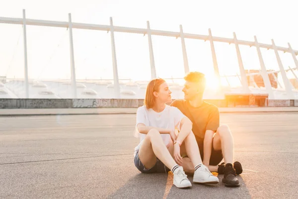 Lyckligt par sitter på gatan i sommarkväll mot bakgrund av solnedgången, ser pojken på flickan och ler. Kärleksfullt par vilar på en kvällspromenad. Livsstilskoncept. — Stockfoto