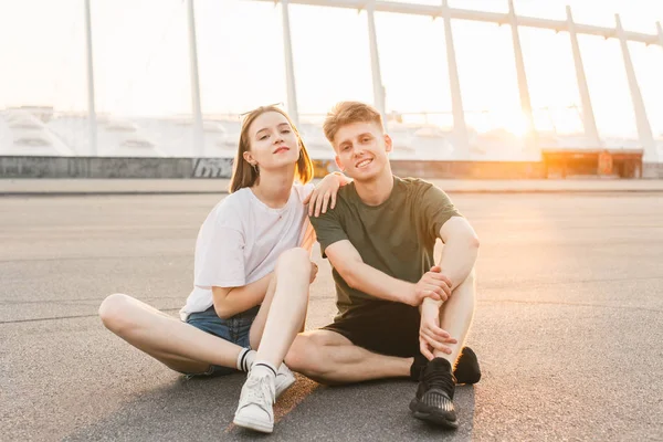 Charming young couple in a stylish casual wear sitting on the background of a beautiful sunset, posing on the camera and smiling.Romantic happy couple relaxing in evening with a beautiful background — стоковое фото