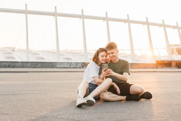 Positieve paar kerel en meisje zitten op straat in de achtergrond van de zonsondergang en gebruiken een smartphone. Stijlvolle liefdevolle paar ontspannen, kijkend naar het scherm van de smartphone en glimlachend. Levensstijl — Stockfoto