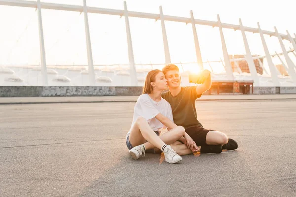 Gelukkig paar ontspannen op de achtergrond van de zonsondergang, meisje kussen jonge man in haar Wang, jongen neemt selfie en glimlach. Stijlvol paar zittend op de achtergrond zonsondergang en moderne architectuur. Levensstijl — Stockfoto