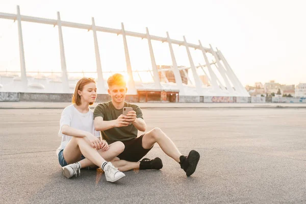 Modernes Paar, das bei Sonnenuntergang auf der Straße im Hintergrund einer wunderschönen Stadtlandschaft sitzt, ein Smartphone benutzt und lächelt. Studenten sitzen auf einem schönen städtischen Hintergrund und entspannen. — Stockfoto
