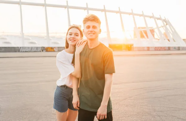 Gelukkig jong stel in Casual kleding poseren tegen de achtergrond van de zon, kijken in de camera en glimlachend. Portret mooie liefhebbers paar, achtergrond van de avond stad en de zonsondergang. Liefdesverhaal — Stockfoto