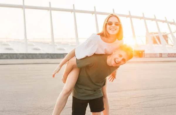 一对微笑的年轻夫妇在夕阳的背景上玩得开心,看着相机的肖像。年轻人背上抱着一个快乐的女孩。美丽的背景上幸福的情侣照片 — 图库照片