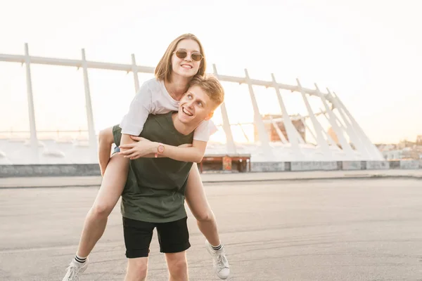 Jonge gelukkige liefhebbers paar in het stedelijke landschap, jonge man houdt het meisje op haar rug. Meisje knuffels de jongen op straat. Stijlvol paar met plezier op de stedelijke achtergrond. Levensstijl — Stockfoto