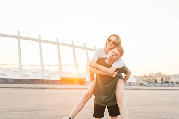 Coppia divertente divertirsi su una passeggiata, sfondo del tramonto.Giovane uomo porta una ragazza sorridente sulla schiena, guardando la camera.Coppia allegra abbracciare e ingannare sullo sfondo della città — Foto Stock