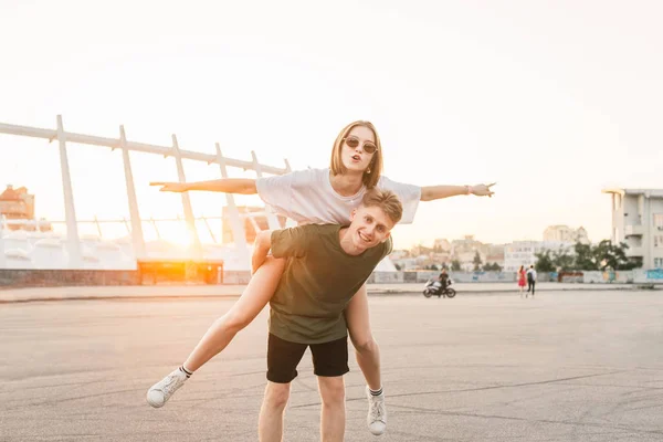 Coppia felice divertirsi al paesaggio della città al tramonto. Attraente ragazza vola sulla schiena di un giovane uomo felice, un giovane uomo sollevato una donna alla moda sulla schiena. Coppia divertirsi in date . — Foto Stock