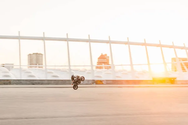 Motociclista fa un trucco pericoloso sullo sfondo del tramonto, cavalcando sulla ruota posteriore.L'uomo guida una moto sullo sfondo di un bellissimo paesaggio della città.Stuntman si allena su una moto . — Foto Stock